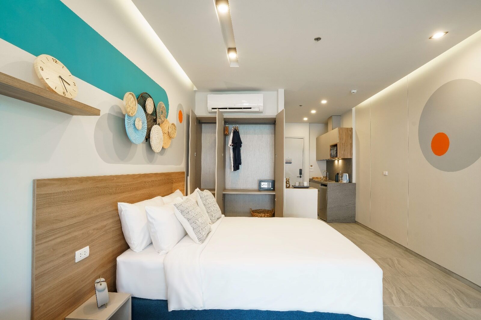Una habitación de hotel moderna con cabecera de madera, una cama grande con ropa blanca, un equipo de aire acondicionado instalado en la pared y una kitchenette al fondo.