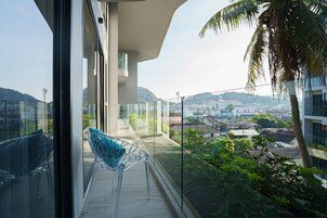 A balcony with clear glass railings, a clear plastic chair, and a view of a cityscape with buildings and hills in the distance.