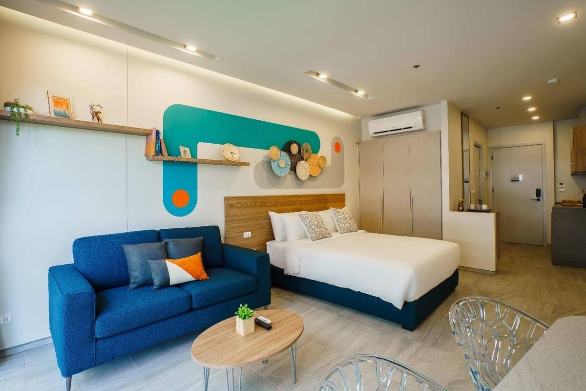 Una habitación de hotel moderna con cama, sofá azul, una mesita y una kitchenette.