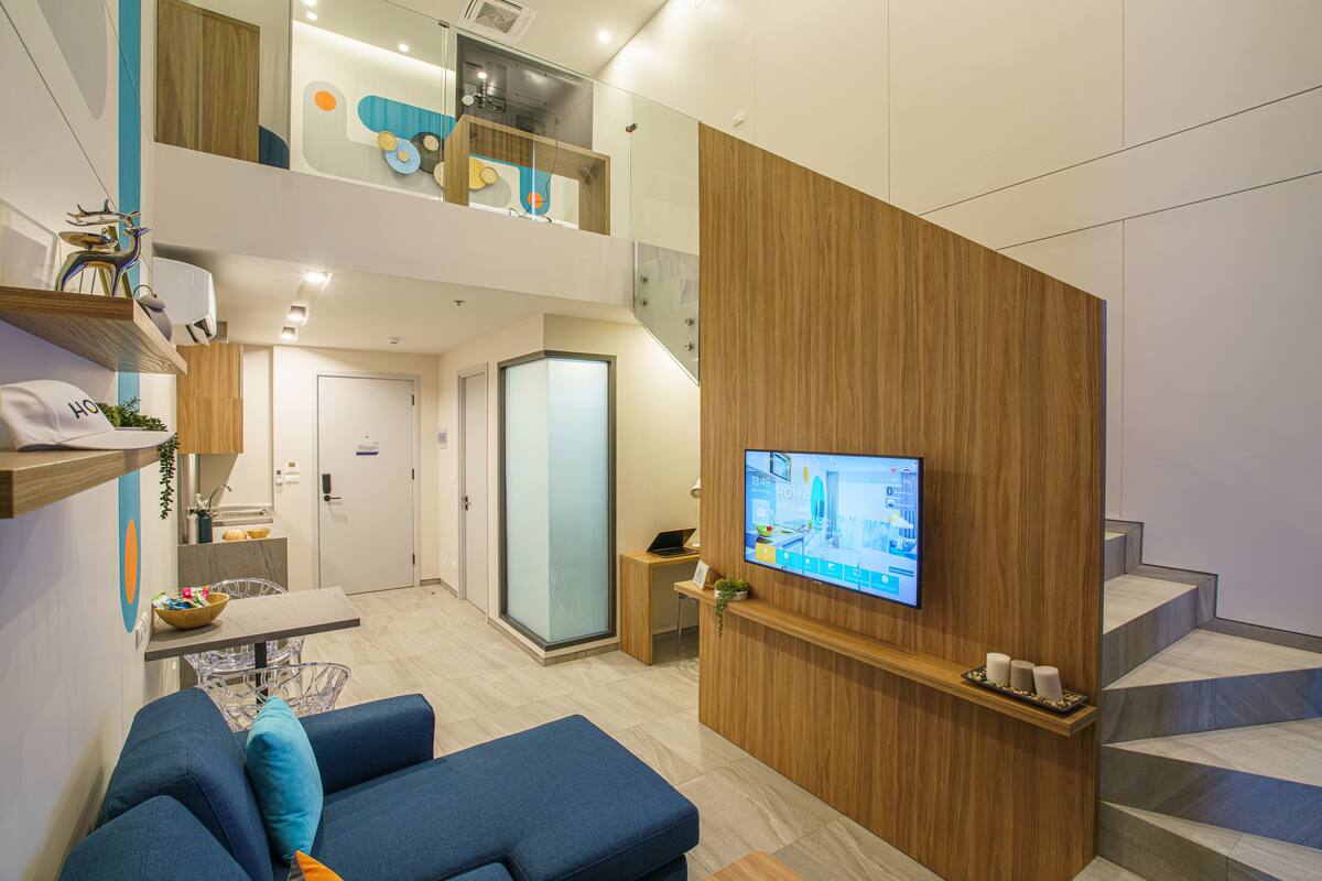 Un espacio de sala moderno con un sofá, un televisor de pantalla plana montado en una pared de madera, una escalera y una pequeña zona de cocina.