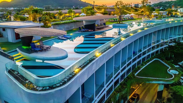 A modern hotel with a large outdoor swimming pool, multiple lounge areas, and a view of a hill in the background. - HOMA Phuket Town (Ratsada)