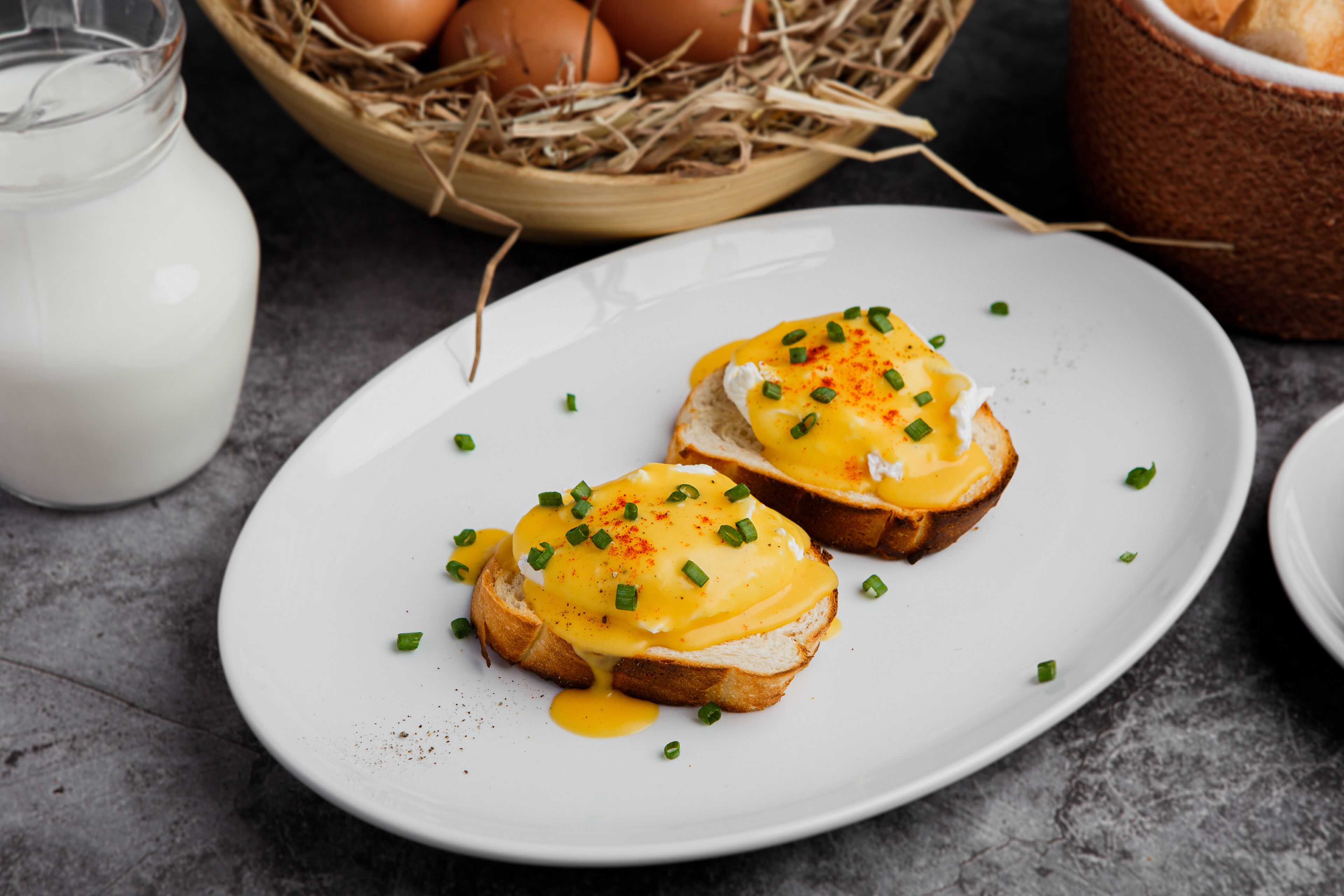 Two pieces of toast with egg and hollandaise sauce on a white plate, with a basket of eggs and a milk jug in the background.