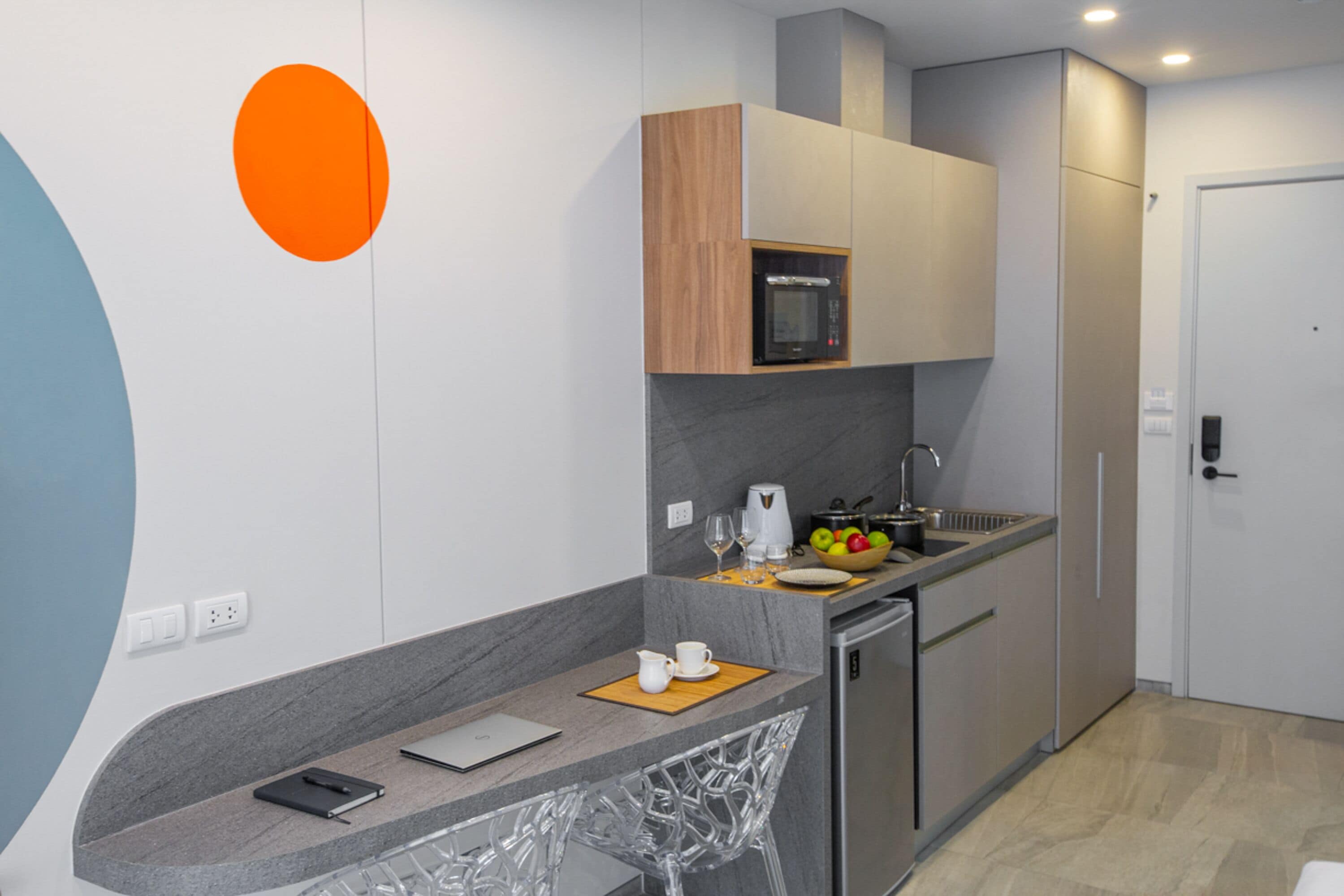 A modern kitchen with a built-in microwave, a sink, and a small refrigerator. There is a wooden cabinet above the microwave and a white door on the right.