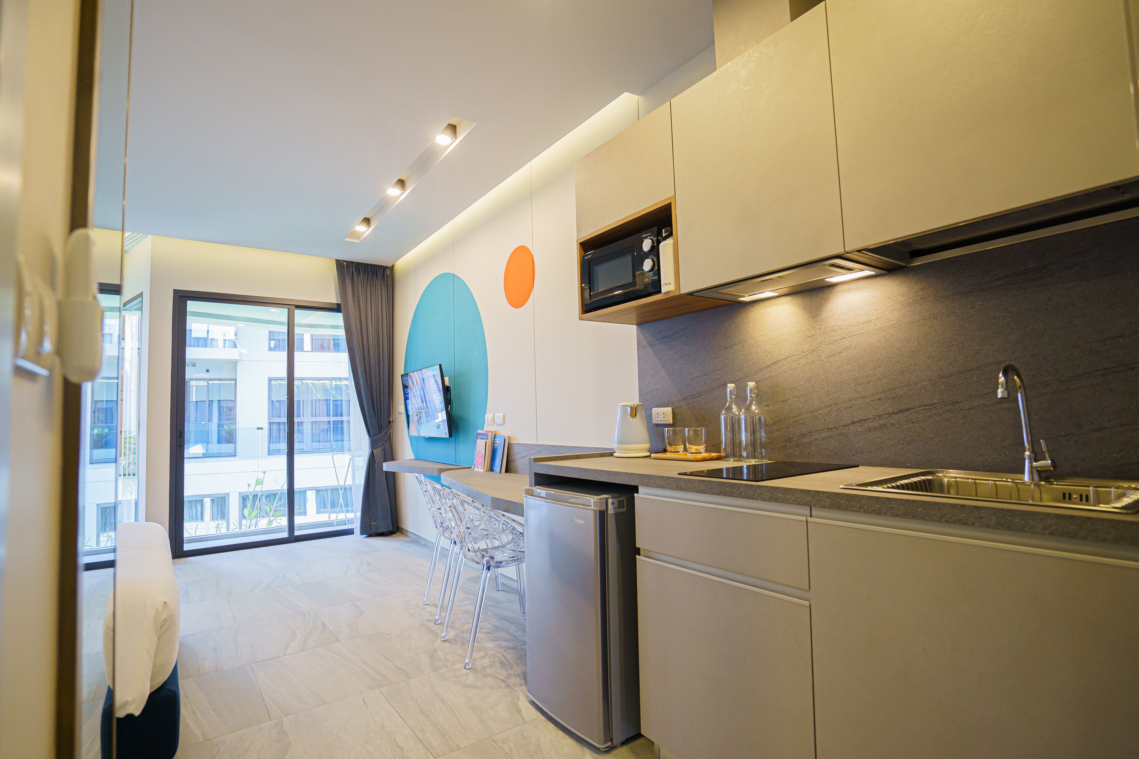 A modern kitchen with a built-in microwave, stainless steel sink, and a refrigerator. There is a bar area with stools and a view of the outside through large windows.