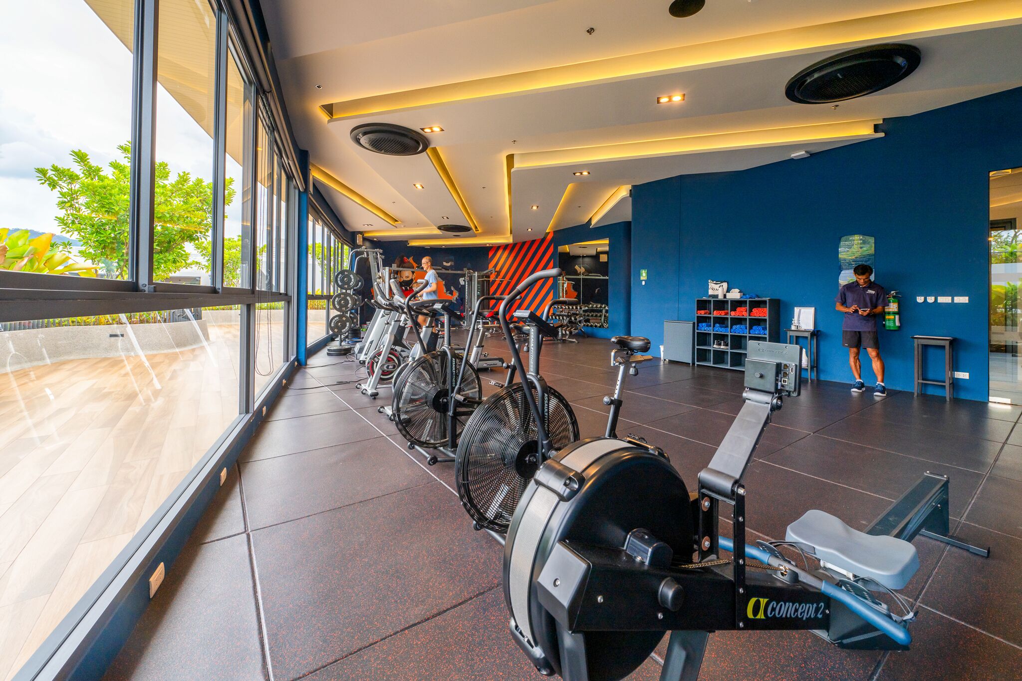 A modern gym with cardio equipment, a reception area, and large windows offering a view of the outdoors.