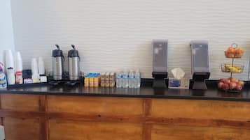 A hotel reception area with a wooden counter, coffee makers, water dispensers, and various beverages and snacks.
