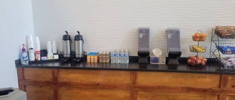 A hotel reception area with a wooden counter, coffee makers, water dispensers, and various beverages and snacks.