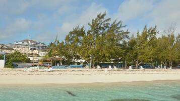 A beach with white lounge chairs and umbrellas, a house in the background, and clear turquoise water.