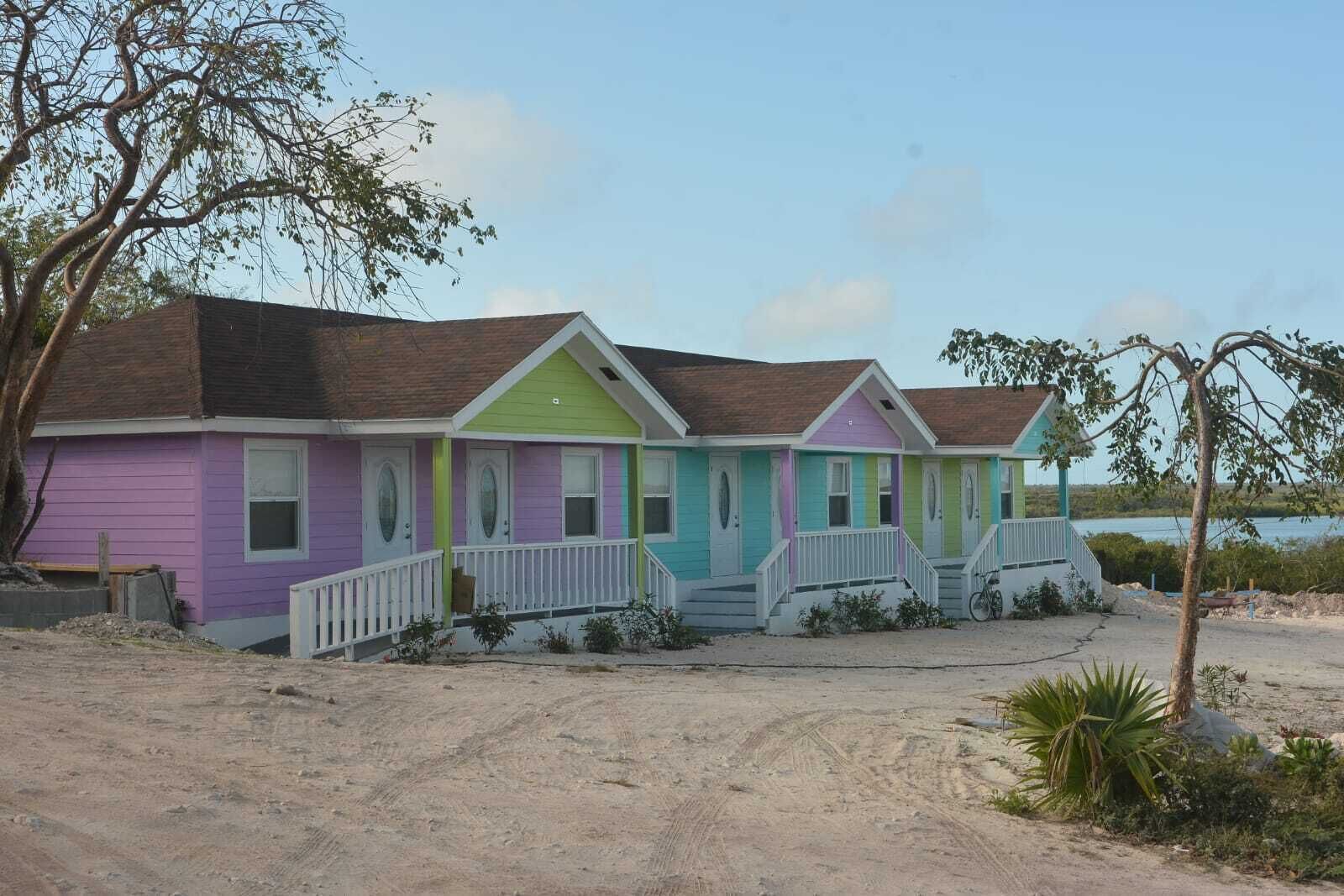 Une rangée de charmantes maisons de plage aux couleurs vives, avec une allée en sable et une vue sur l’eau.