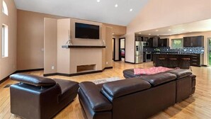 A spacious living area with a large sectional sofa, a flat-screen TV mounted on a wall, and a kitchen area with dark countertops and bar stools. - Spearfish South Dakota rally rental (Spearfish)