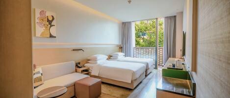 A modern hotel room with a sofa, two beds, a small table, and a view of greenery outside.