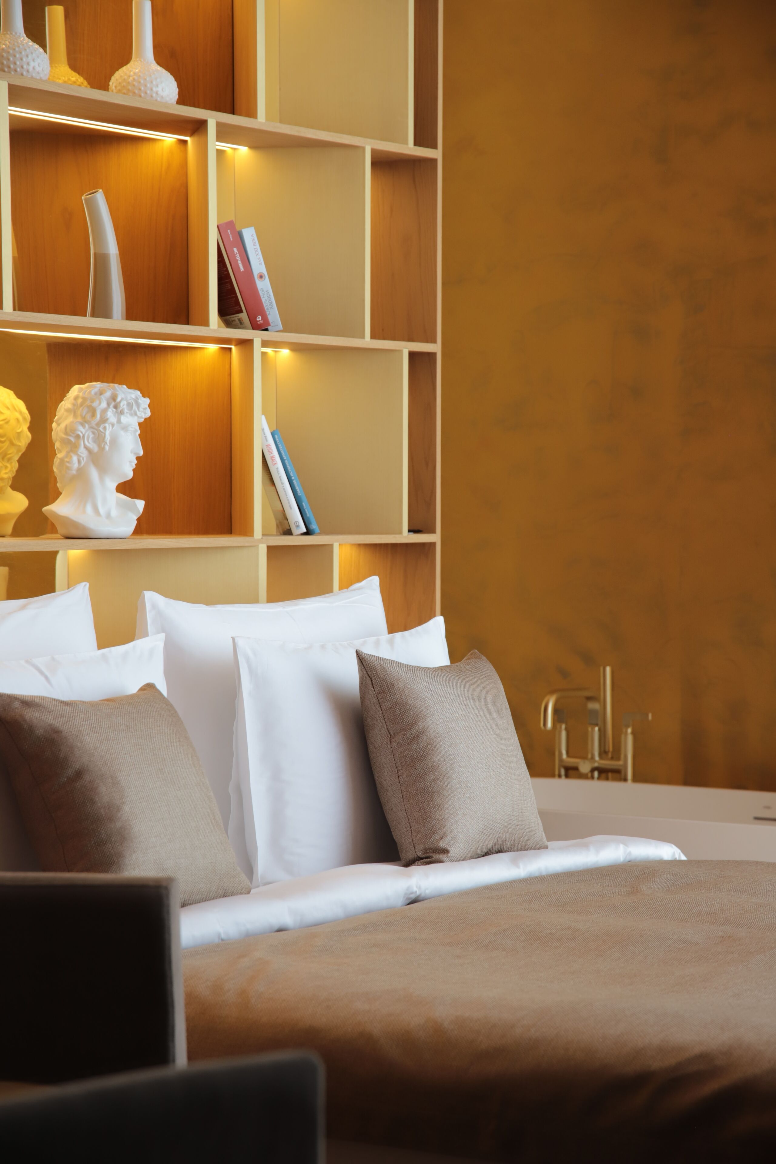 A neatly arranged bedroom with a bed, pillows, a bookshelf, and a bust sculpture.