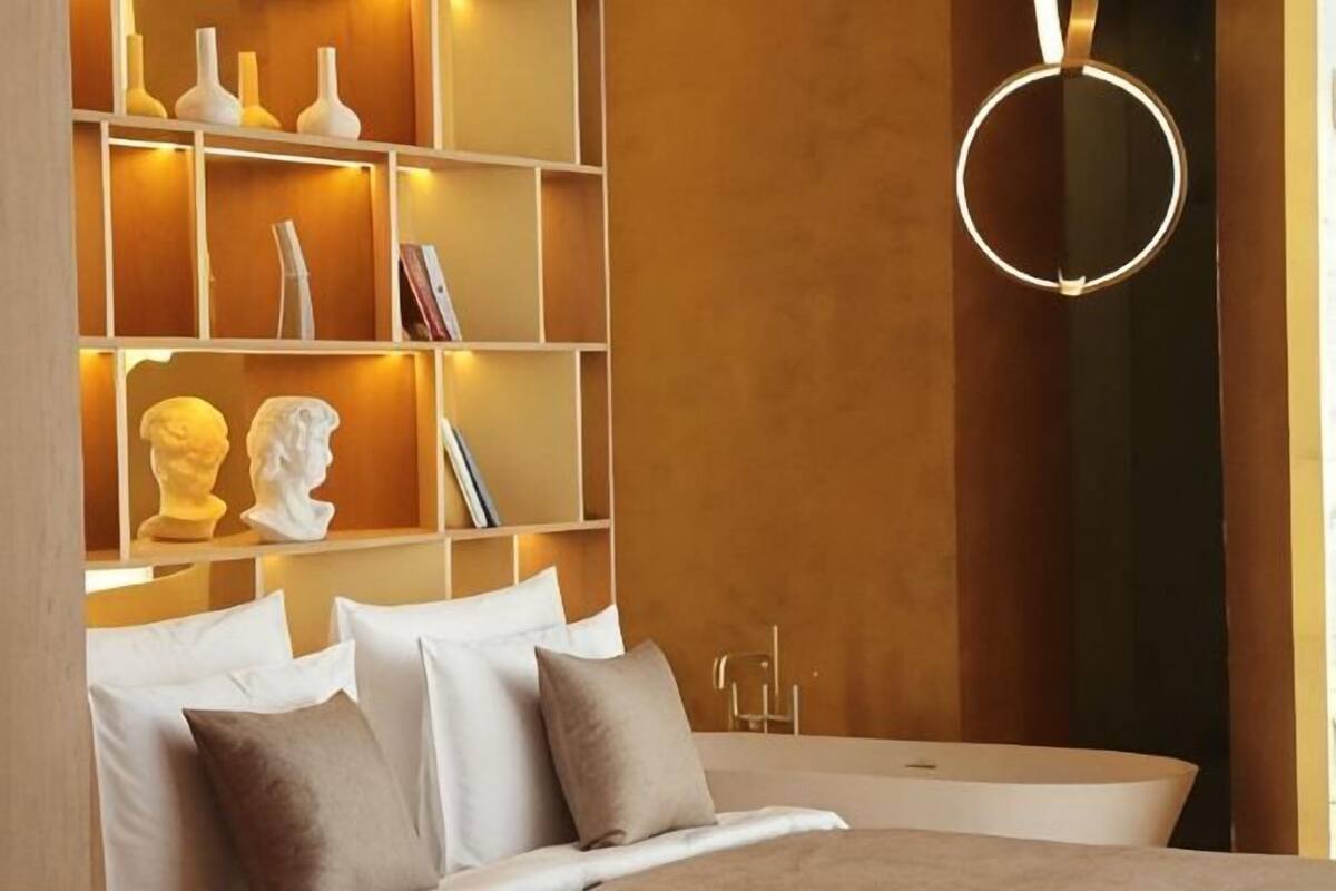 A bedroom with a bed, a bedside table, a wall-mounted shelf with decorative items, and a circular light fixture.