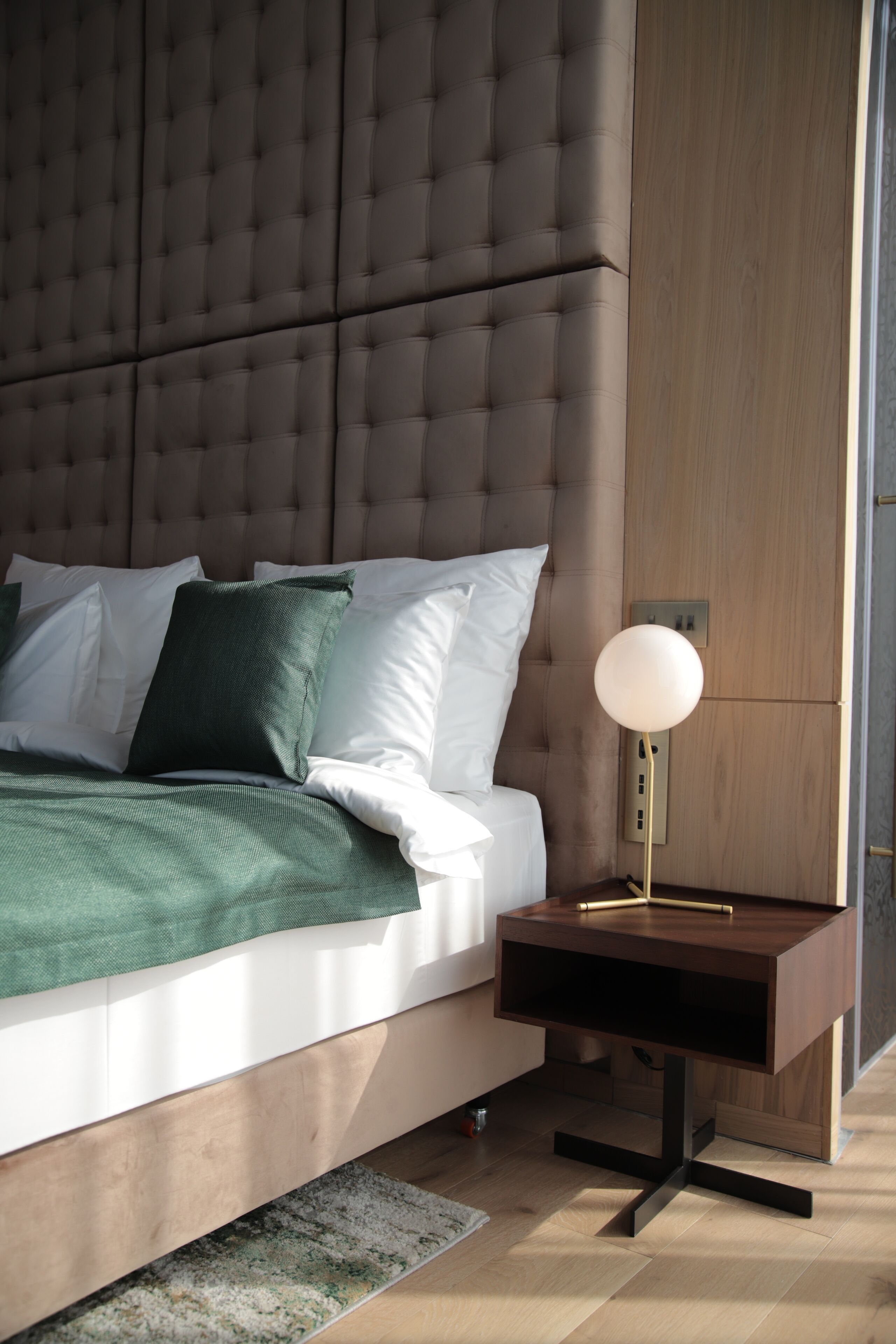 Un dormitorio moderno con una cabecera capitonada, una mesita de noche de madera con una lámpara y una manta verde.