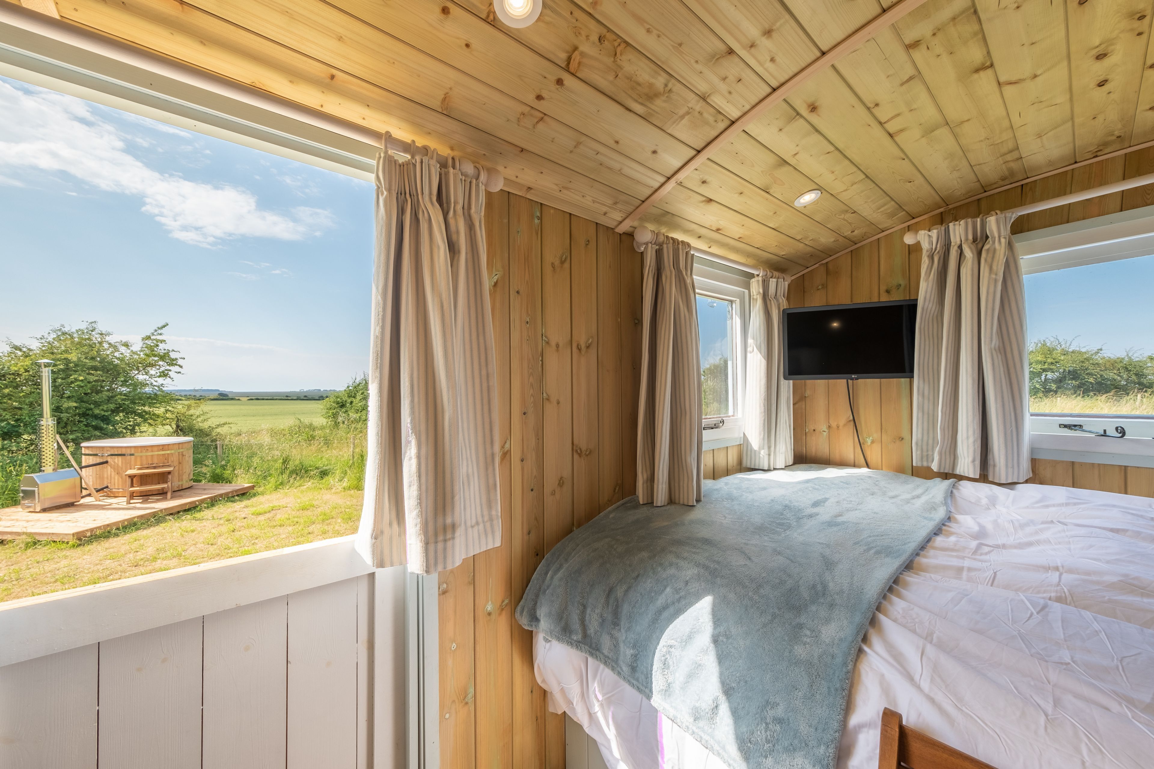 A wooden cabin bedroom with a bed, curtains, a TV, and a view of the outdoors.