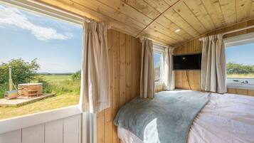 A wooden cabin bedroom with a bed, curtains, a TV, and a view of the outdoors.