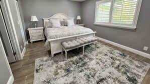A bedroom with a bed, nightstands, a bench, and a large window with blinds. - Beautiful Cottage located inside PGA National (Palm Beach Gardens)