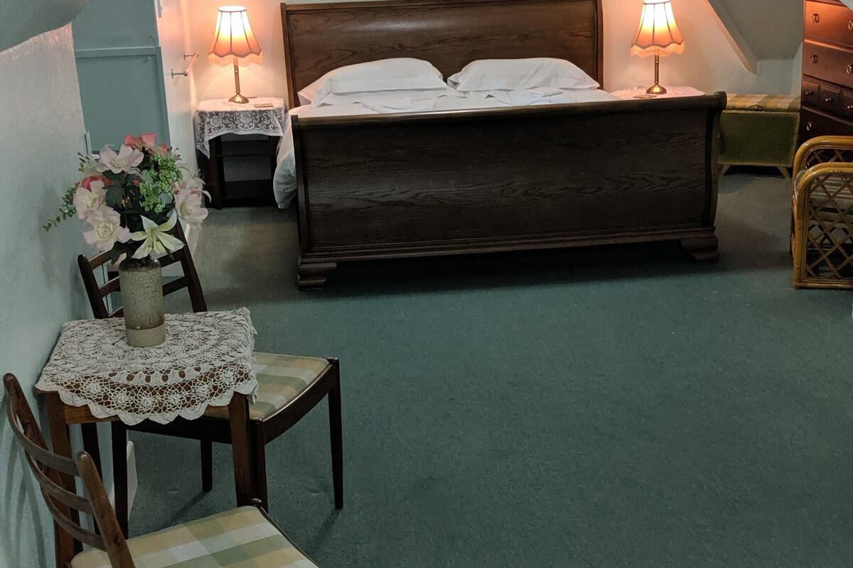 A cozy attic bedroom with a large bed, two bedside lamps, a wooden chair, and a small table with a vase of flowers.