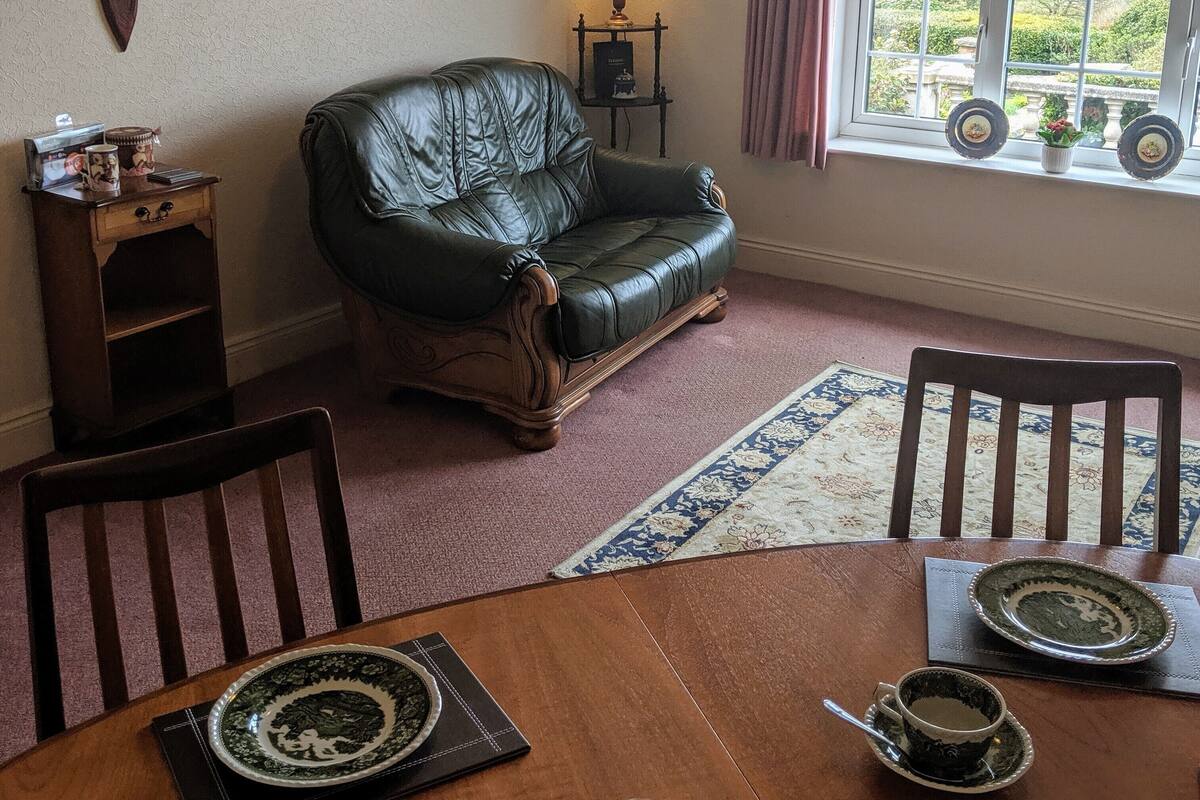 A cozy living room with a leather sofa, a wooden side table, a lamp, and a view through a window.