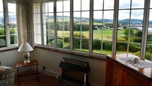 A room with large windows offering a view of rolling hills and a river. There is a piano and a lamp on a side table.
