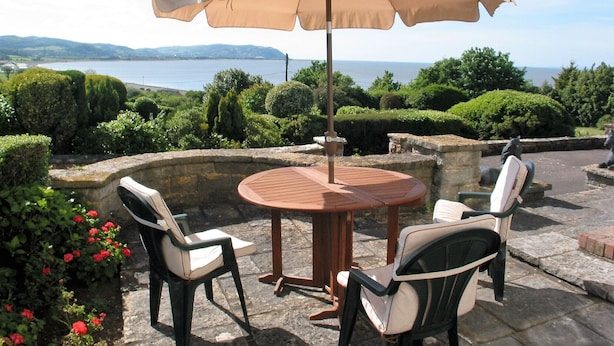 A patio with a round wooden table and two chairs, an umbrella, and a view of the sea and hills.