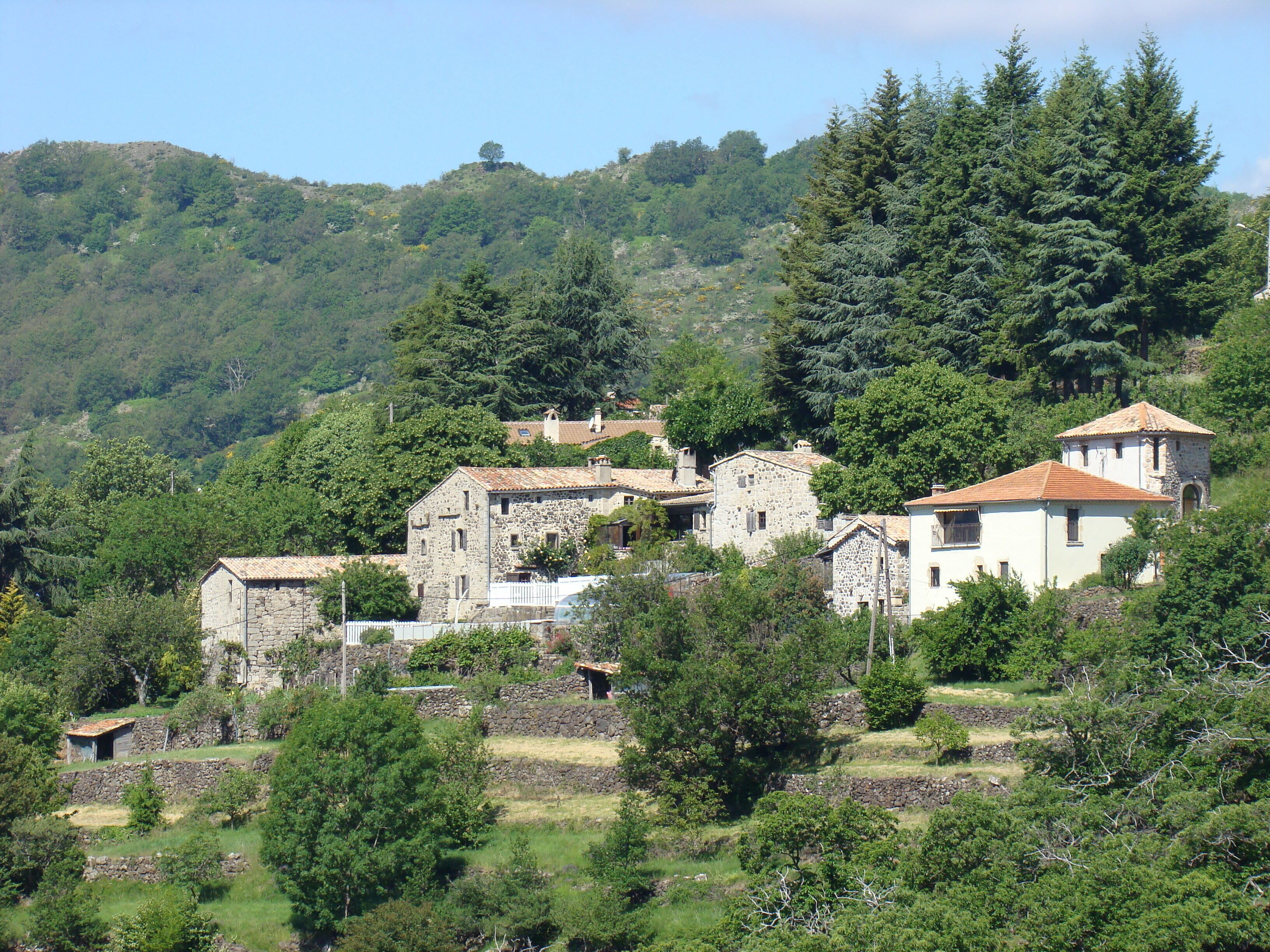 Un village perché sur une colline, avec des bâtiments en pierre, dont certains sont coiffés de toits en tuiles rouges, le tout entouré d’une végétation luxuriante et d’arbres.