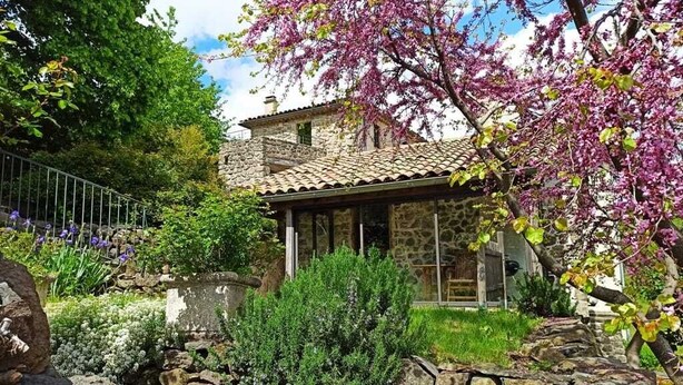 Una casa de piedra con tejado de baldosas, rodeada por un jardín con plantas en flor y un muro de piedra.