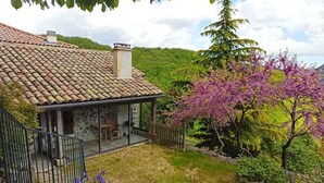 Una casa de piedra con techo de tejas, un patio cubierto y un jardín con flores moradas.