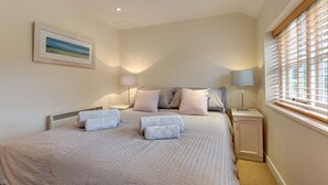 Une chambre à coucher comprenant un lit, deux tables de chevet, une commode, une lampe, une fenêtre avec des stores et un tableau encadré accroché au mur.