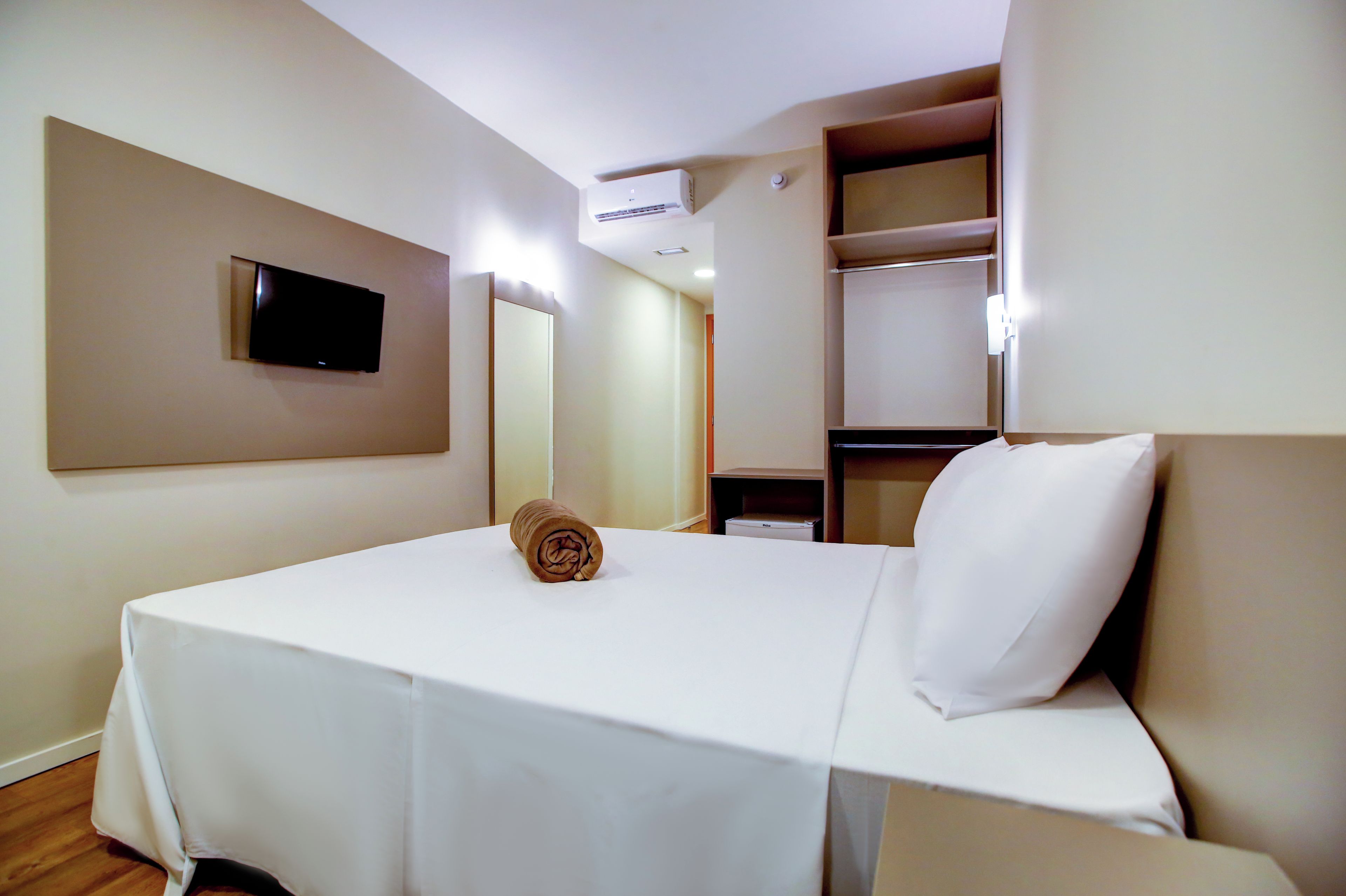 A hotel room with a bed, a TV mounted on the wall, a mirror, and a shelf with a few items.
