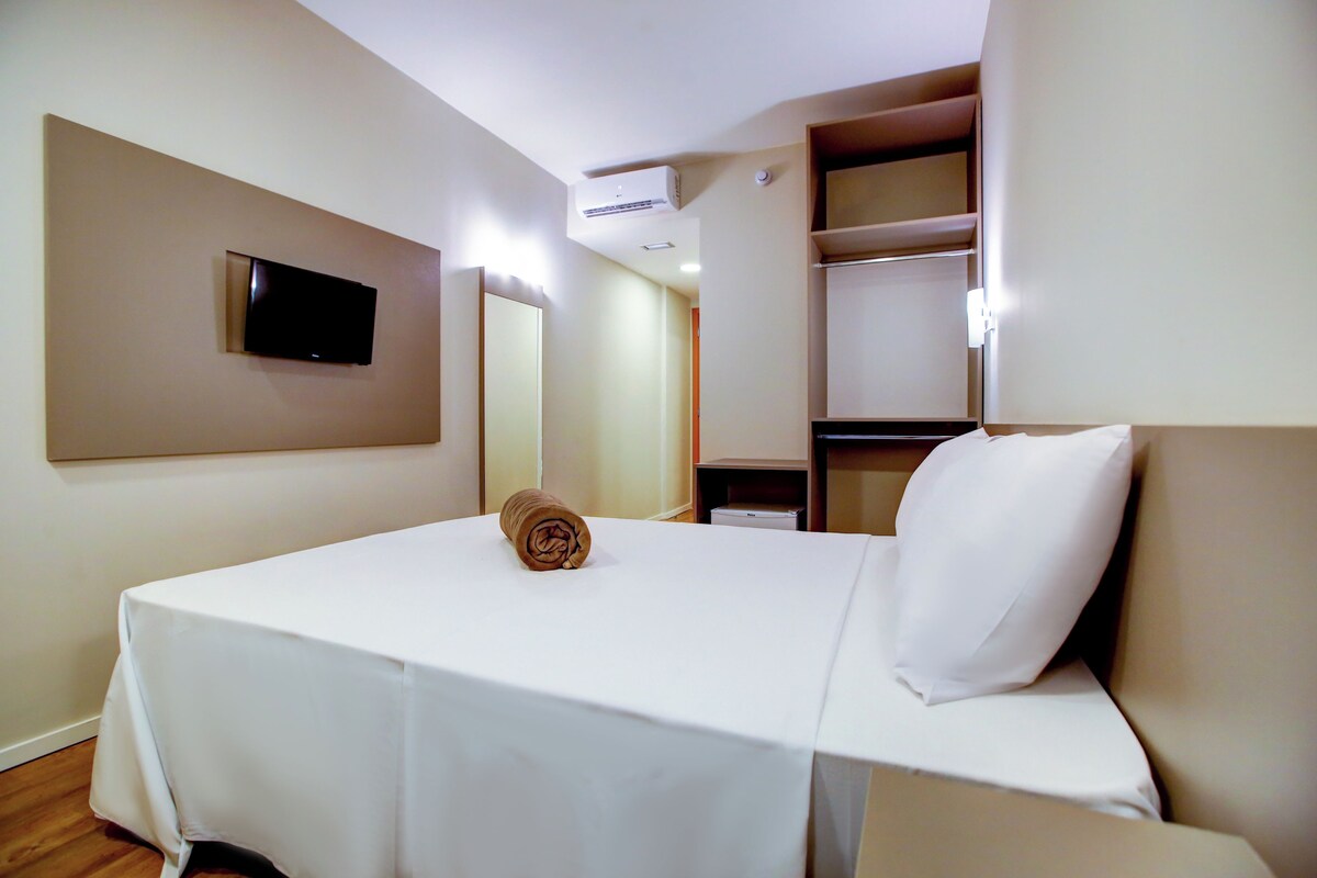 Habitación de hotel con cama, televisor montado en la pared, espejo y estante con algunos objetos.