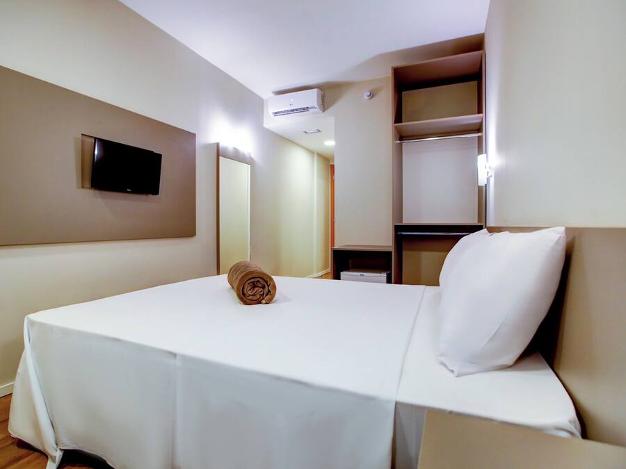 Een hotelkamer met een bed, een aan de muur bevestigde televisie, een spiegel en een plank met enkele spullen.