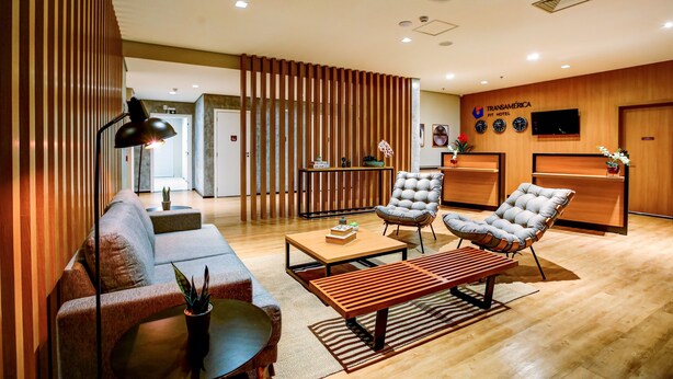 A modern hotel lobby with a wooden reception desk, a sofa, two armchairs, a coffee table, and a lamp. - Transamerica Fit Lucas Do Rio Verde (Lucas do Rio Verde)