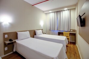 Habitación de hotel con dos camas individuales, un escritorio, una silla, un televisor montado en la pared y una ventana con cortinas.