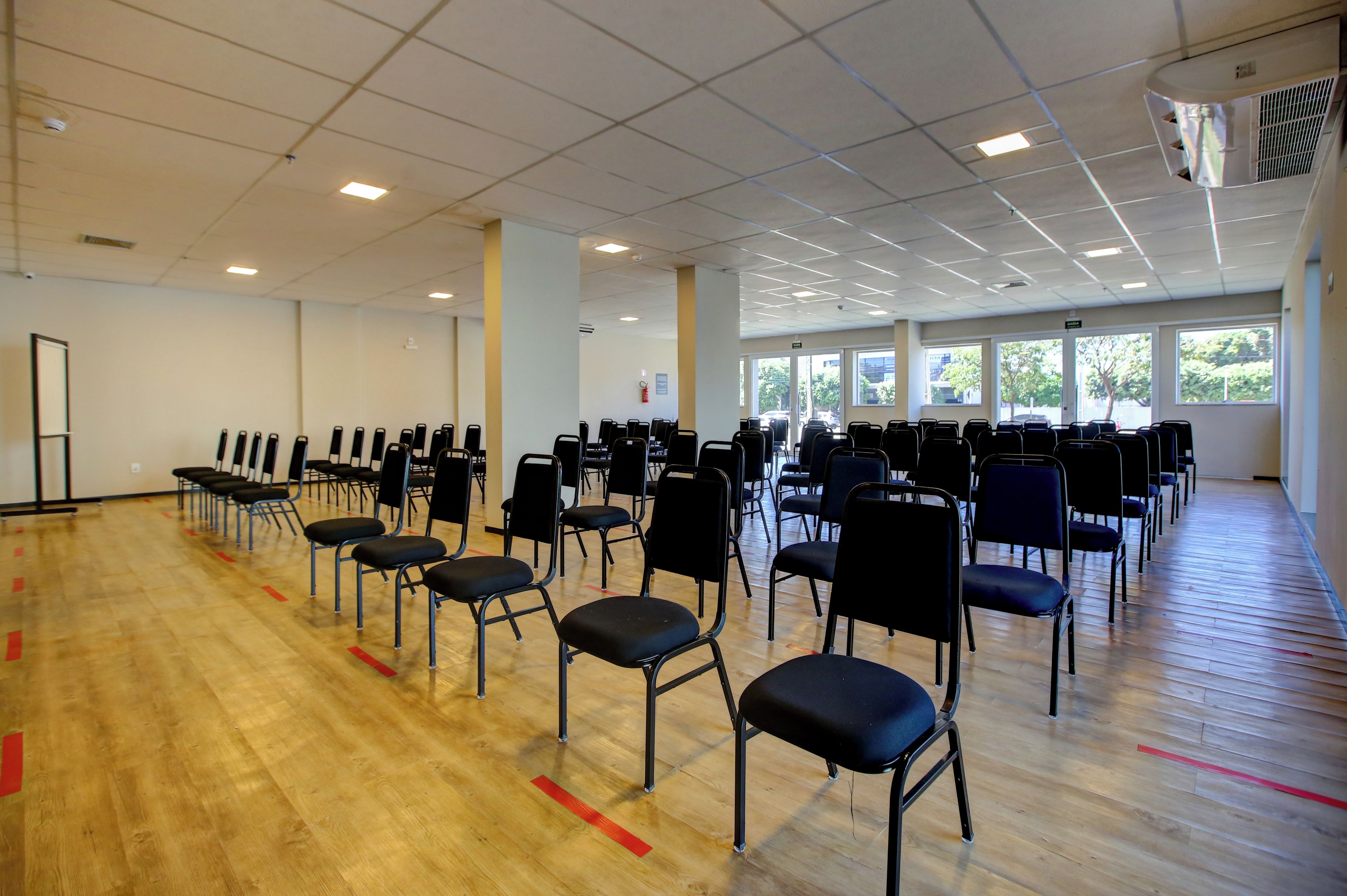 A large indoor space with rows of black chairs arranged for seating, a wooden floor, and a ceiling with fluorescent lighting.