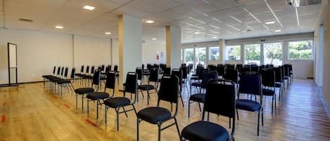 A large indoor space with rows of black chairs arranged for seating, a wooden floor, and a ceiling with fluorescent lighting.