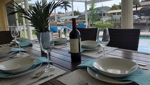 Une table dressée pour le dîner, avec une bouteille de vin, des verres et des assiettes, donnant sur une piscine et un espace de détente extérieur.