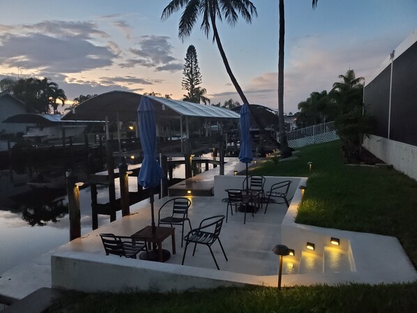 An outdoor patio with tables and chairs, a covered seating area, and a view of the water and palm trees.