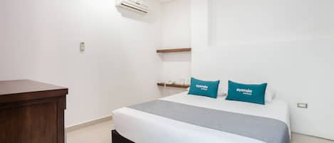 A hotel room with a bed, a wooden desk, an air conditioner, and a shelf with a phone and a cup.