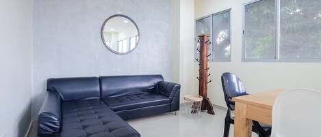 Una sala de estar moderna con un sofá seccional azul oscuro, un espejo redondo, una mesa de comedor de madera con dos sillas y un mueble alto de madera.