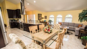 A spacious kitchen with dark cabinets, a dining area with a glass table and wicker chairs, and a living room with a sofa and a large plant. - Palm Tree III Pool Home Waterfront  (Apollo Beach)