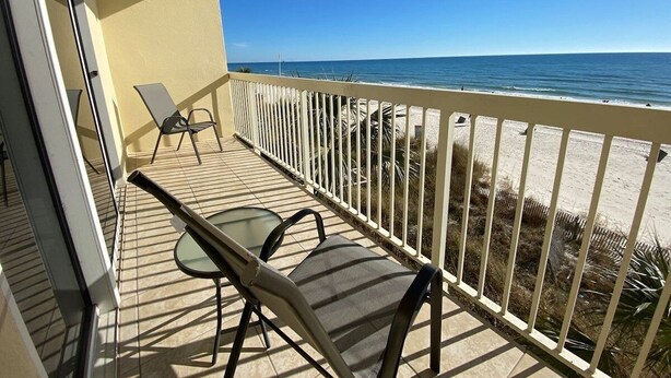 A balcony with a table and chairs, overlooking a beach with a fence and vegetation.