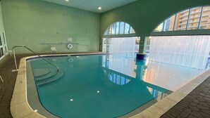 An indoor swimming pool with a curved edge, a tiled floor, and a window with a view of buildings.