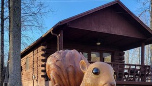 Een groot houten beeld van een eekhoorn die een eikel vasthoudt, geplaatst op een ronde basis met een boomstam. Op de achtergrond staat een houten chalet met een veranda.