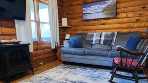 Ein gemütliches Blockhaus-Interieur mit einer Holzwand, einem grauen Sofa, einem Schaukelstuhl, einem schwarzen Holzofen und einem großen Fenster mit Vorhängen.