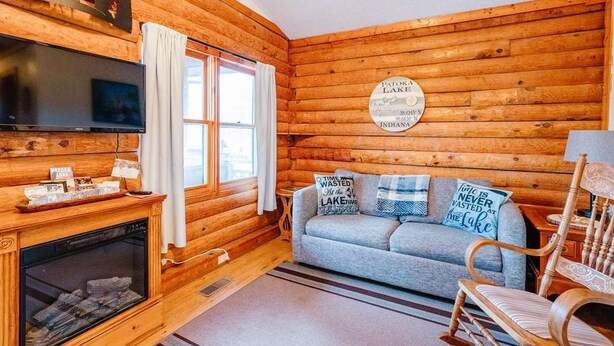 A cozy cabin interior with a fireplace, a sofa with decorative pillows, a television, and a wooden wall with a circular plaque. - Log Cabin 4 at Osborn Boat Ramp on Patoka Lake (Jackson Township)