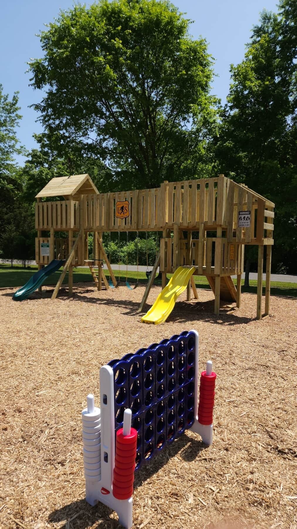 A wooden play structure with a slide and a climbing wall, surrounded by trees.