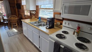A compact kitchen with white cabinets, a stove, an oven, a coffee maker, and a window with blinds. - Log Cabin 9 at Osborn Boat Ramp on Patoka Lake (Jackson Township)