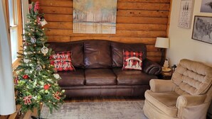 Et hyggeligt sommerhusinteriør med en trævæg, en brun lædersofa, en beige lænestol, et juletræ og et billede af en skov.