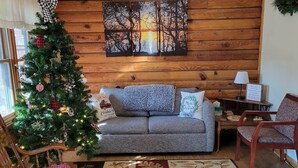 Una sala de estar acogedora con una pared de madera, un árbol de Navidad, un sofá gris, un sillón mecededor y una alfombra estampada.
